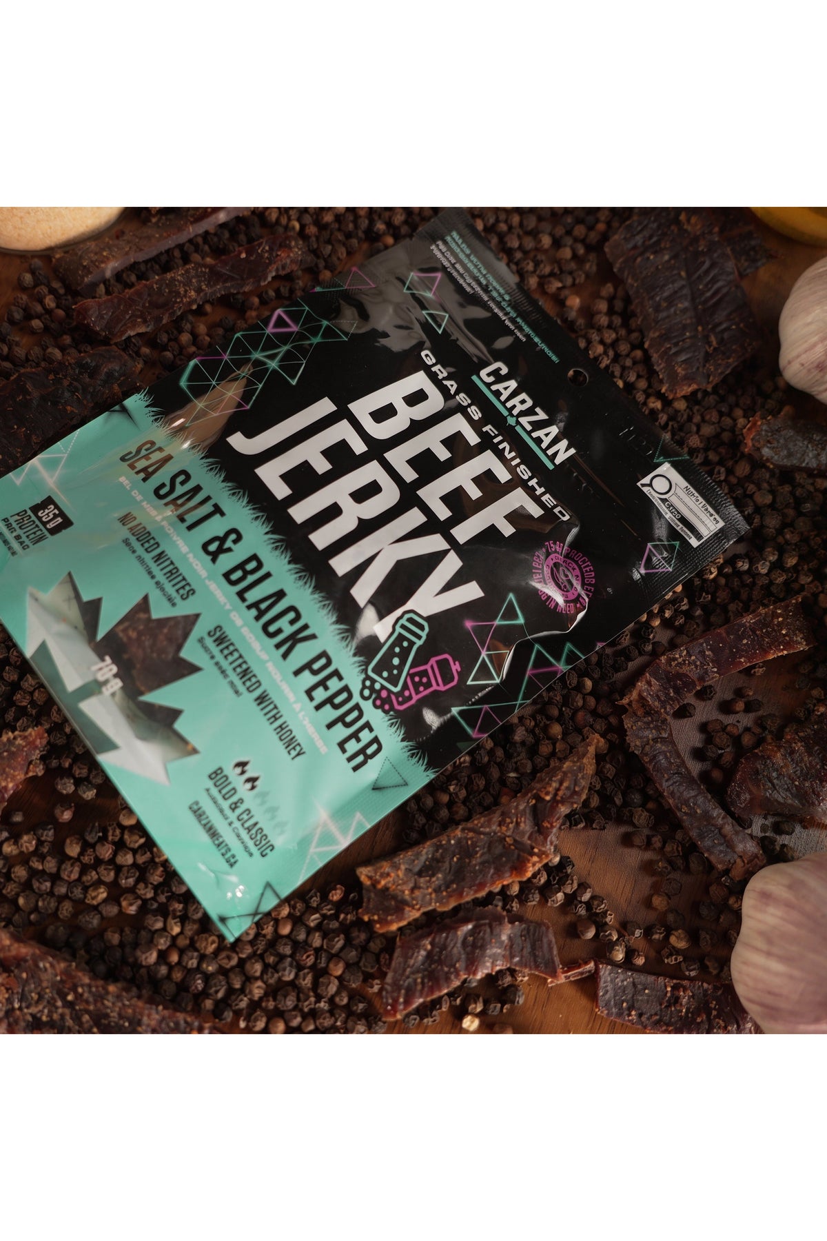Carzan Sea Salt Black Pepper 70g beef jerky bag surrounded by peppercorns, garlic and onion, with loose jerky pieces on a wood table