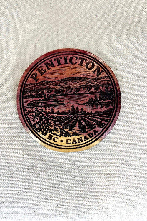 Close-up of the Two Makers Crew Penticton cedar magnet showing the engraved lake, hills, steamship and vineyard design.