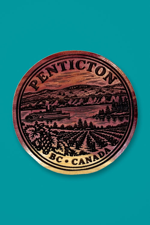 Two Makers Crew Penticton cedar magnet with engraved Okanagan scenery against a teal background.